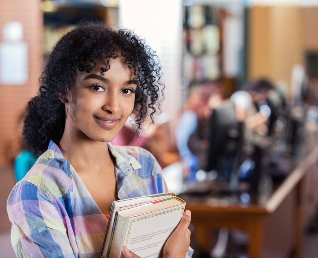 Teen or young adult African American woman with curly natural hair is smiling and looking at the camera while standing in modern college library. College student is holding textbooks and is standing in front of station with computers and bookshelves.